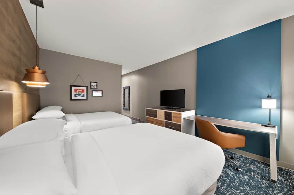 Contemporary hotel bedroom with two queen beds, blue accent wall, orange pendant light, desk, and neutral tones