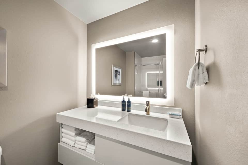 Modern bathroom vanity with white countertop, illuminated mirror, and shower visible