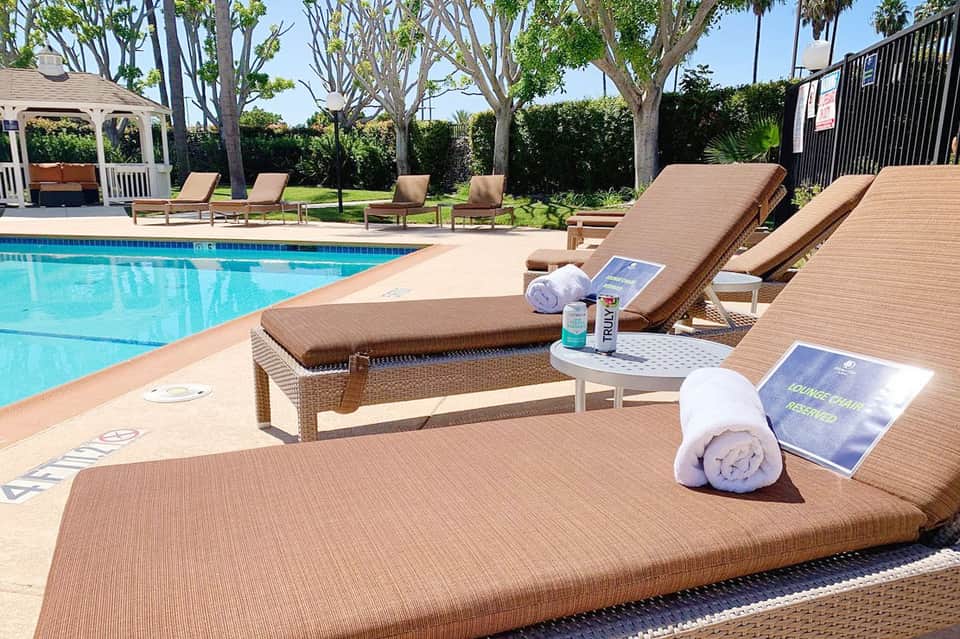 Resort pool with brown lounge chairs, rolled towels, and refreshments on side tables near palm trees