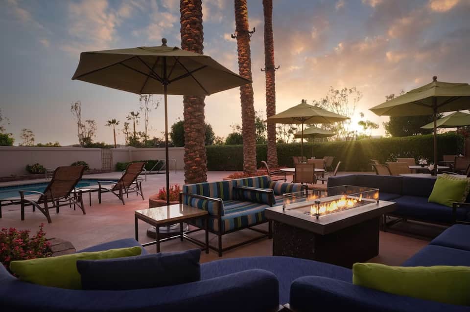 Resort patio at sunset with fire pit, striped seating, lounge chairs, umbrellas, pool, and tall palm trees