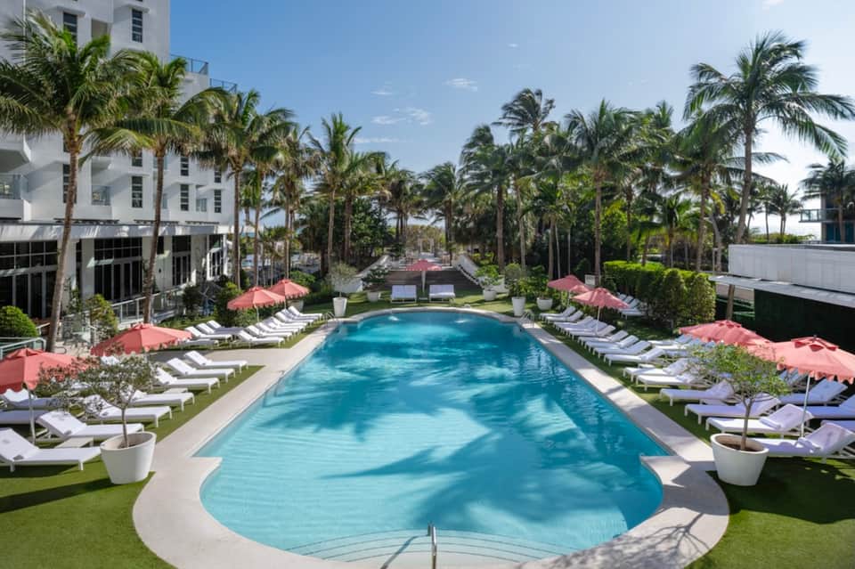 Rectangular resort pool surrounded by palm trees, lounge chairs, pink umbrellas, and white buildings