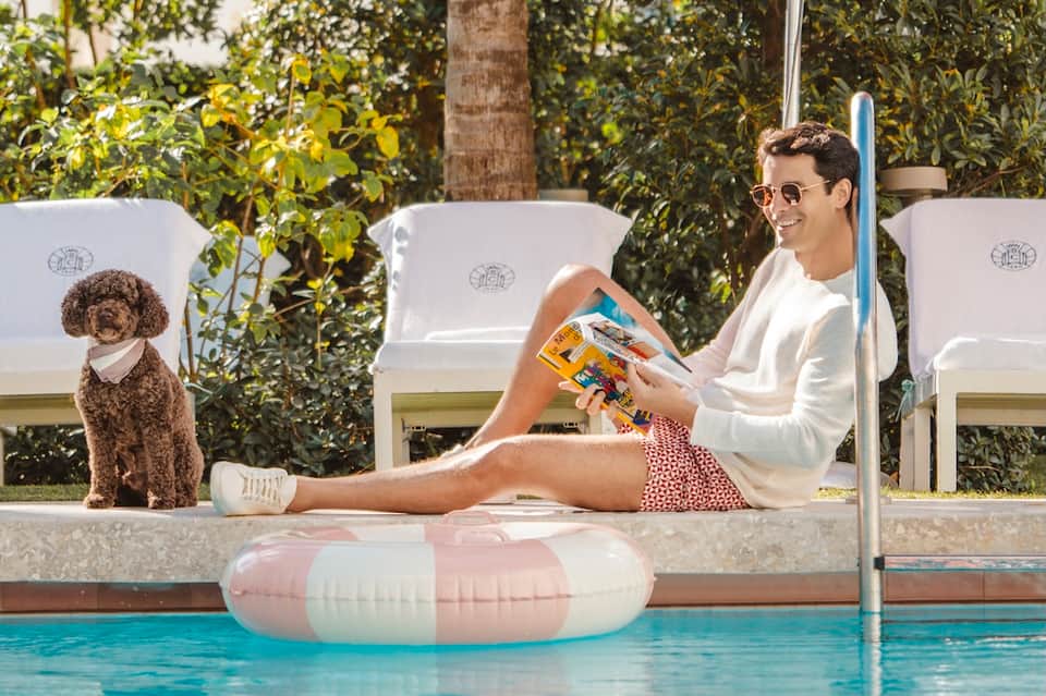 Man relaxing by pool reading magazine with pet dog nearby and lounge chairs under shade