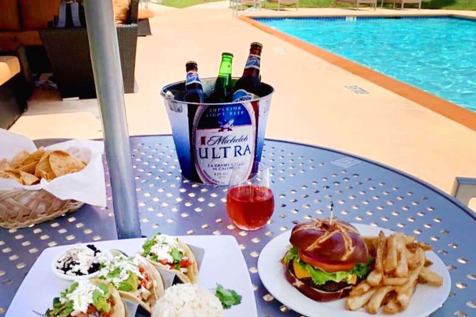 Poolside dining table with beer bucket, tacos, burger, fries, and tropical setting