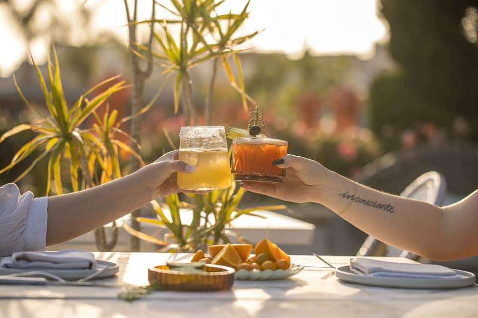 Two hands toasting with cocktails at sunset dining table with tropical plants and flowers