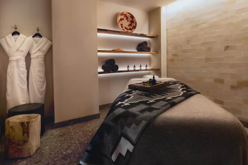 Spa treatment room with massage table, white spa robes, wooden shelves with amenities, and neutral stone tiling