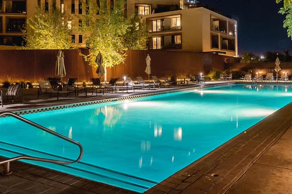 Nighttime resort pool with turquoise water, illuminated umbrella seating, lush landscaping, and modern building architecture