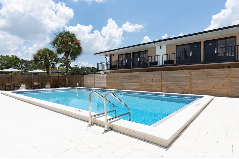 Rectangular swimming pool with white deck, metal railings, palm trees, wooden fence, and two-story building with balconies