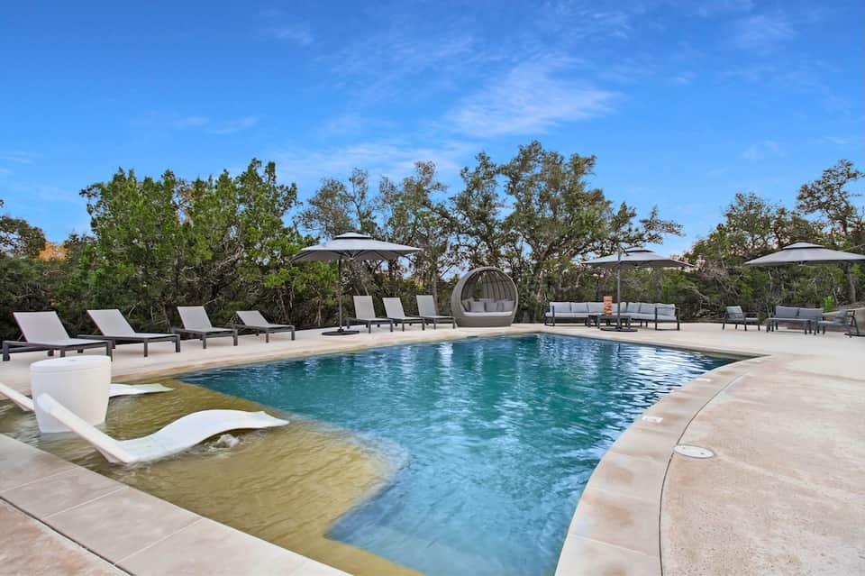 Resort infinity pool with shallow lounge area, white loungers, umbrellas, and natural landscaping
