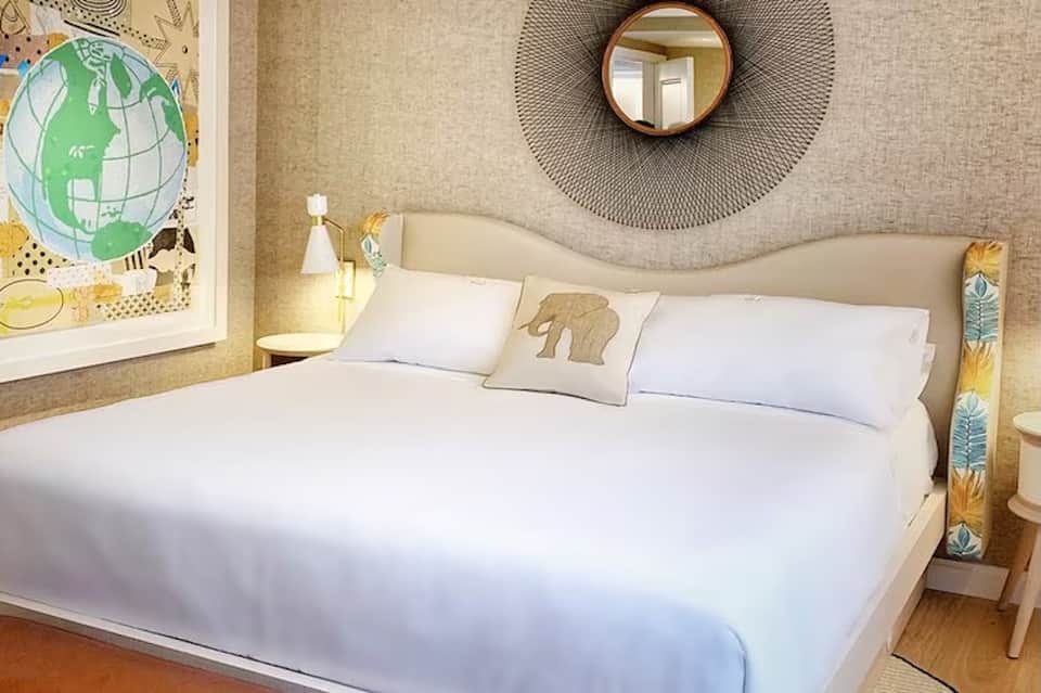 Modern hotel bedroom with cream upholstered headboard, circular wall mirror, white bedding, and wood accents