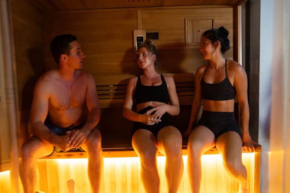 Sauna interior with wooden walls and benches, three people in athletic wear relaxing with warm amber lighting