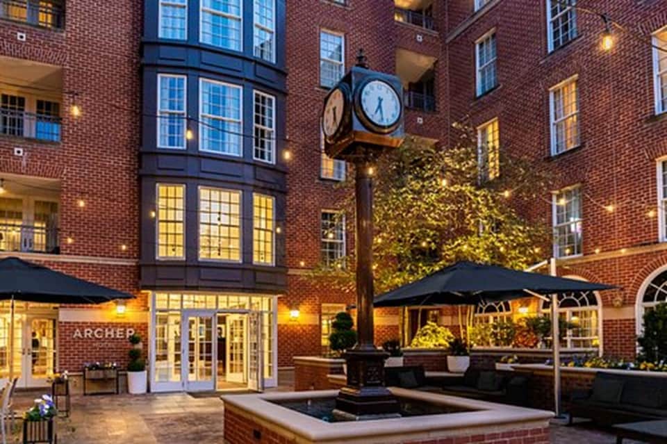 Brick courtyard at dusk with illuminated storefronts, clock, umbrellas, and fountain