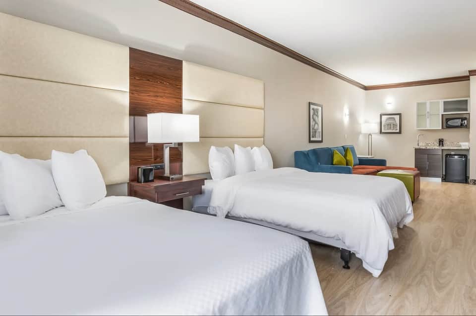 Hotel suite with two queen beds, cream headboards, sitting area, kitchenette, and modern furnishings