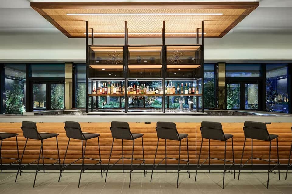 Modern hotel bar with wooden counter seating, illuminated liquor shelves, and contemporary design elements