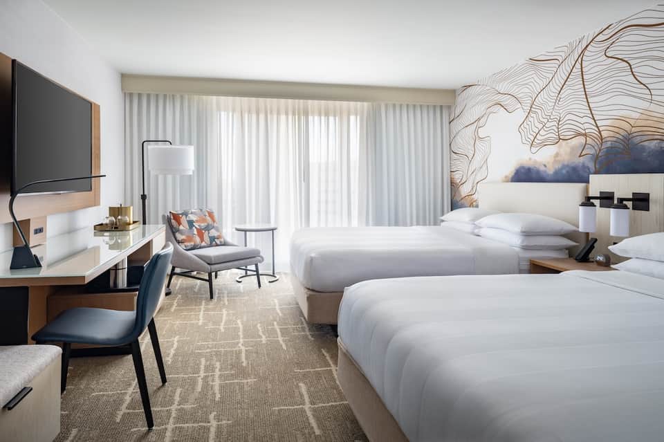 Contemporary hotel room with two queen beds, wall-mounted TV, desk, and floor-to-ceiling windows