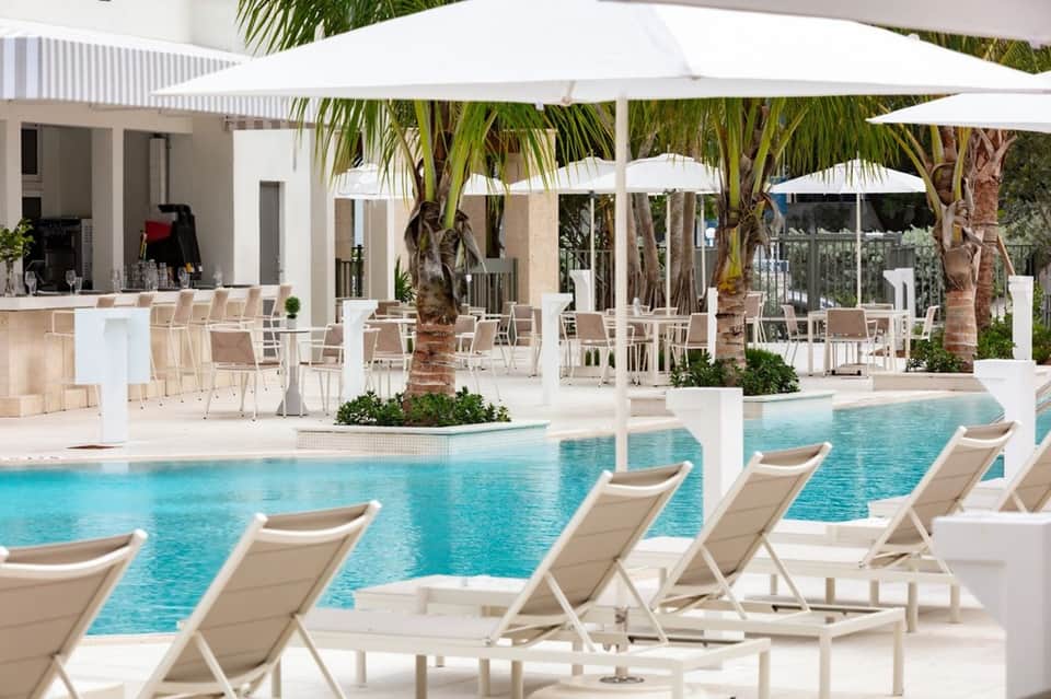 Luxury resort pool area with white loungers, umbrellas, bar counter, and palm trees
