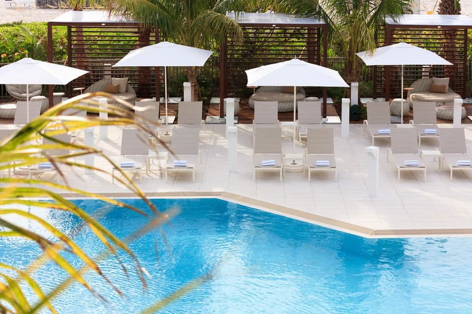 Resort pool deck with white loungers, umbrellas, wooden privacy screens, and tropical setting