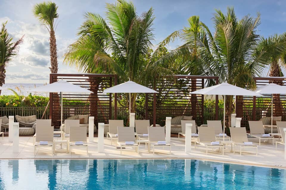 Beachfront resort pool deck with palm trees, white umbrellas, lounge chairs, and ocean horizon backdrop