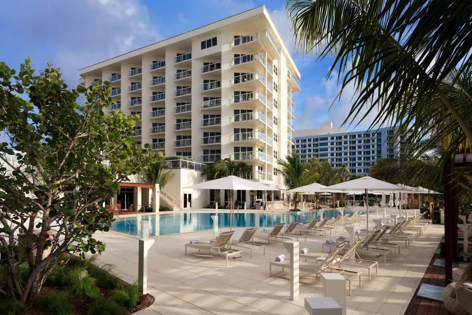 Resort pool surrounded by lounge chairs, white umbrellas, modern building, and palm trees