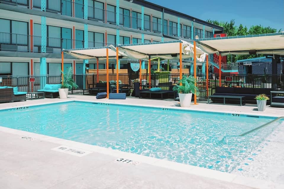 Outdoor pool with white shade sails, colorful umbrellas, and multi-story hotel building