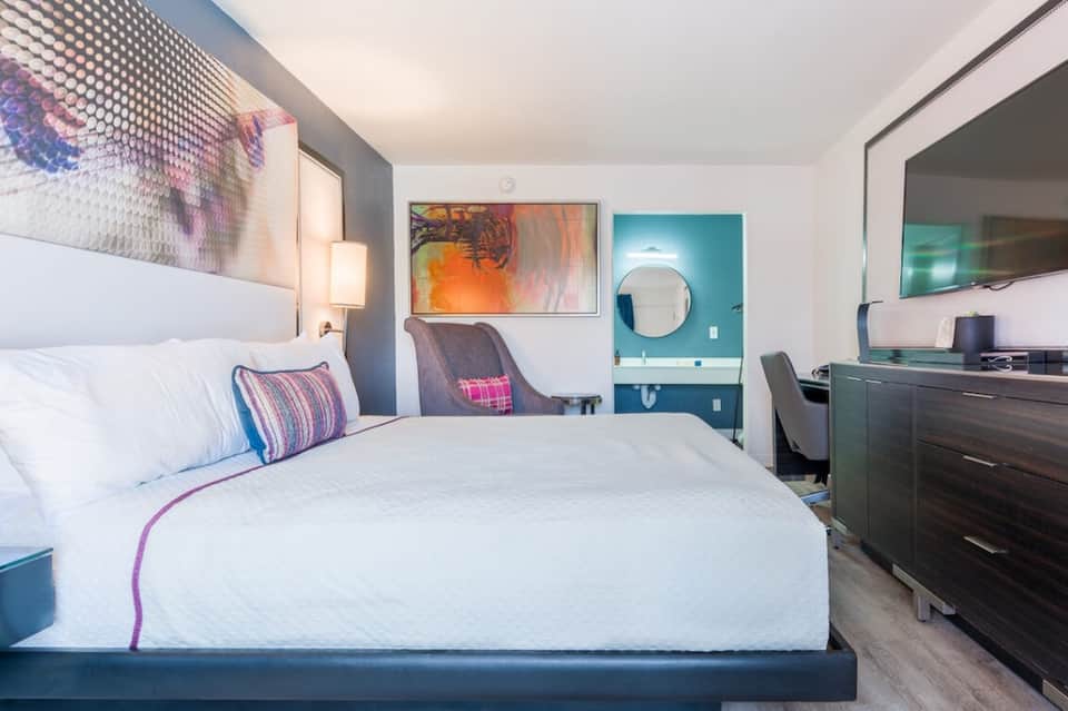 Modern hotel bedroom with queen bed, colorful accents, and teal bathroom entrance