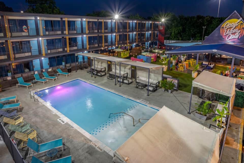 Vibrant nighttime pool scene with colorful lighting, hotel rooms, and entertainment area