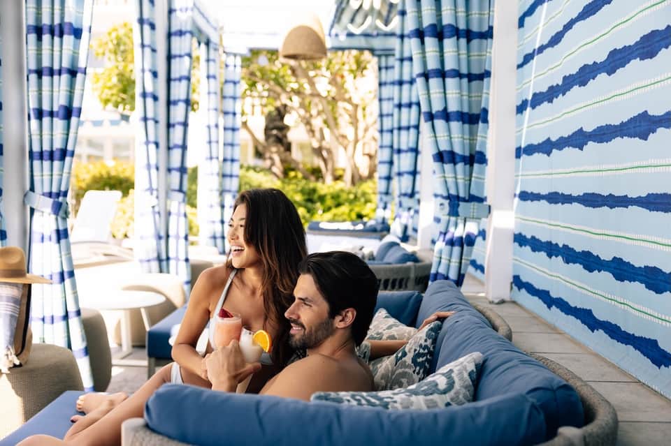 Couple relaxing in a private cabana with blue and white striped curtains overlooking the pool area