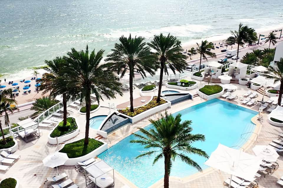 Beachfront resort pool with palm trees, white lounge chairs, and ocean view with sandy beach