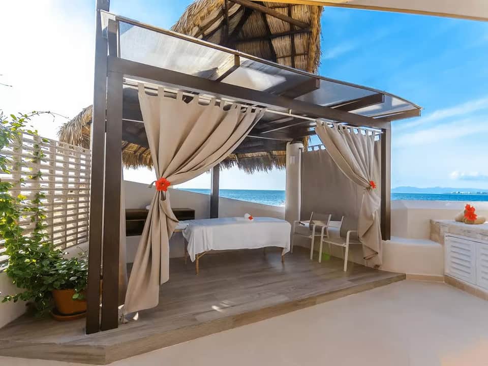 Beachfront cabana with white pergola, draped curtains, ocean view, decorative flowers, and lounge table