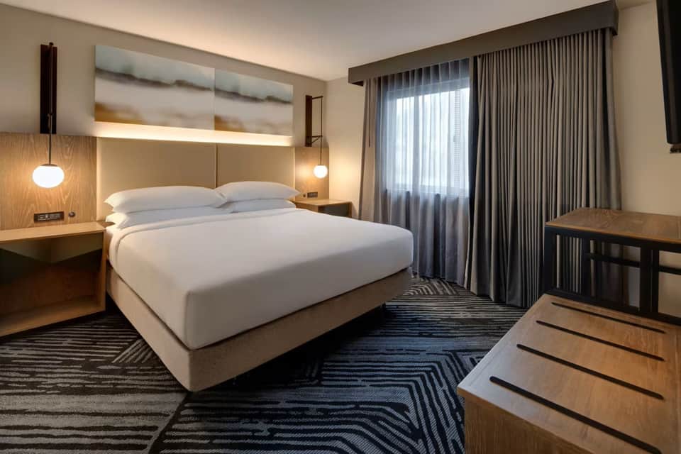 Modern hotel bedroom featuring queen bed, wood accents, pendant lights, and floor-to-ceiling windows