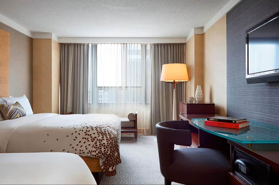 Hotel room with queen bed, desk area, dark brown upholstered chair, sheer curtains, and warm neutral color palette