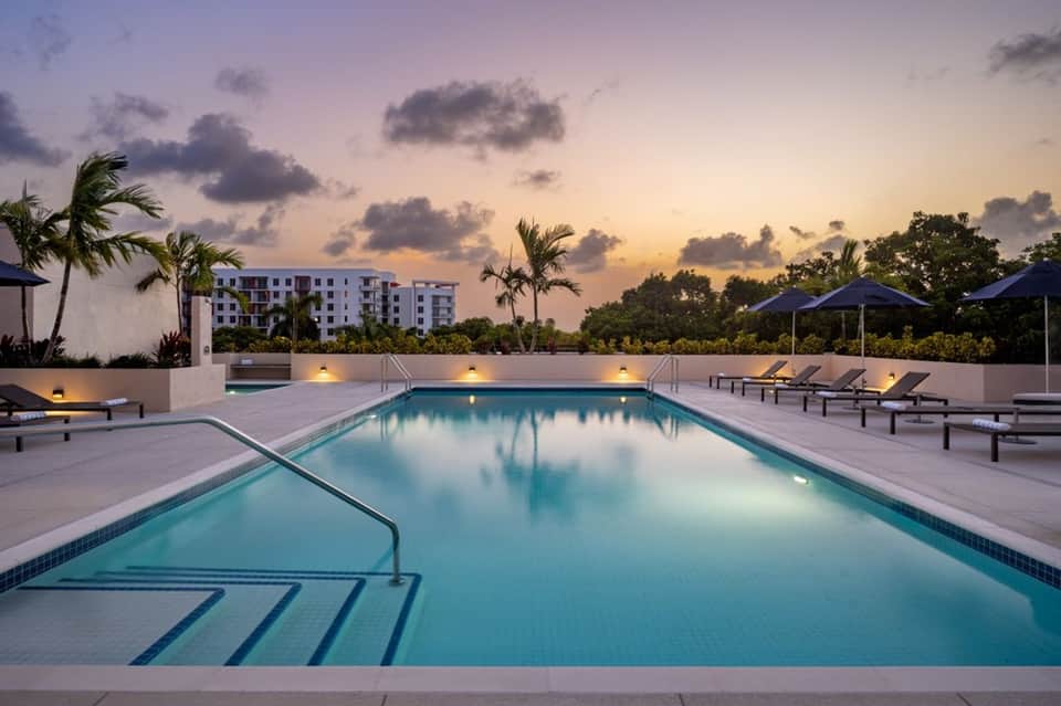 Resort pool at twilight with lounge chairs, umbrellas, illuminated deck, and residential building backdrop
