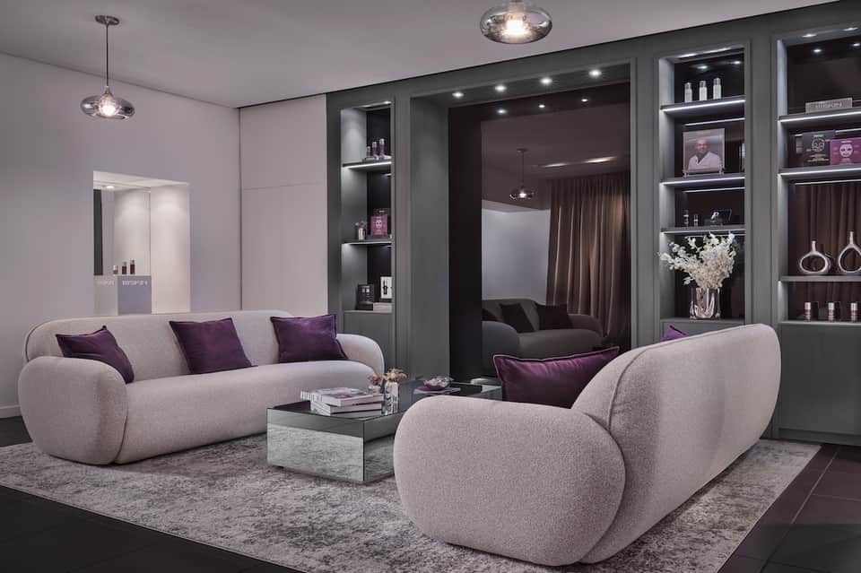 Luxury spa lounge with curved beige seating, dark shelving with beauty products, and soft pendant lighting