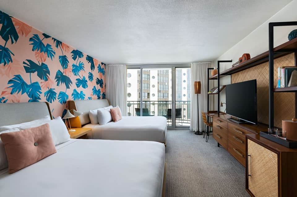 Hotel bedroom with twin beds, tropical leaf wallpaper, balcony doors, and entertainment center