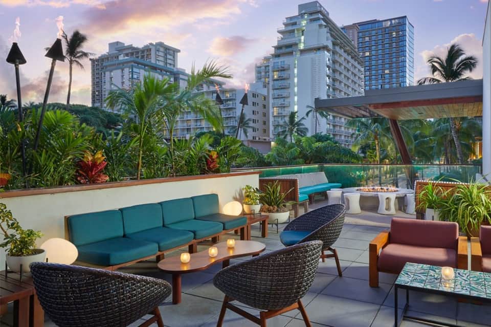 Rooftop lounge at dusk with teal sofa, rattan chairs, tropical plants, and city skyline