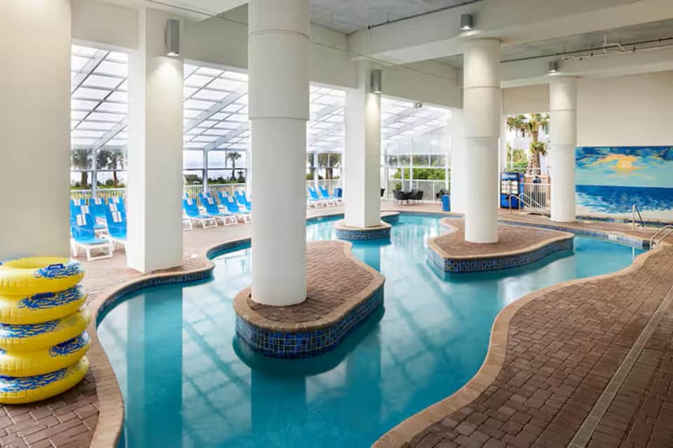Indoor resort pool with bright skylights, multiple pools, loungers, and white support columns