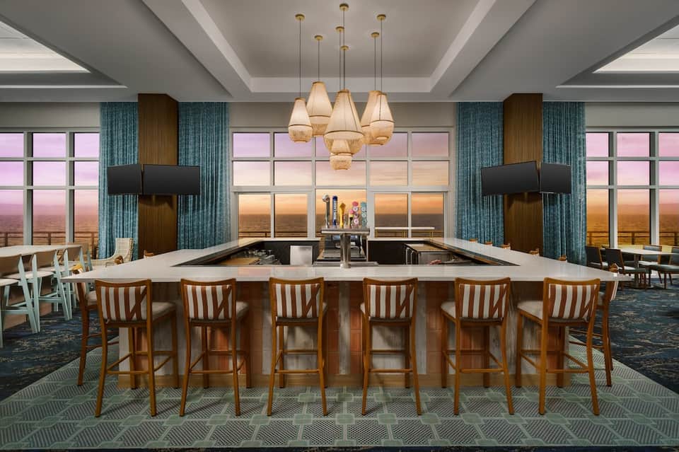Modern rooftop bar with expansive windows, fireplace, pendant lights, and sunset ocean view