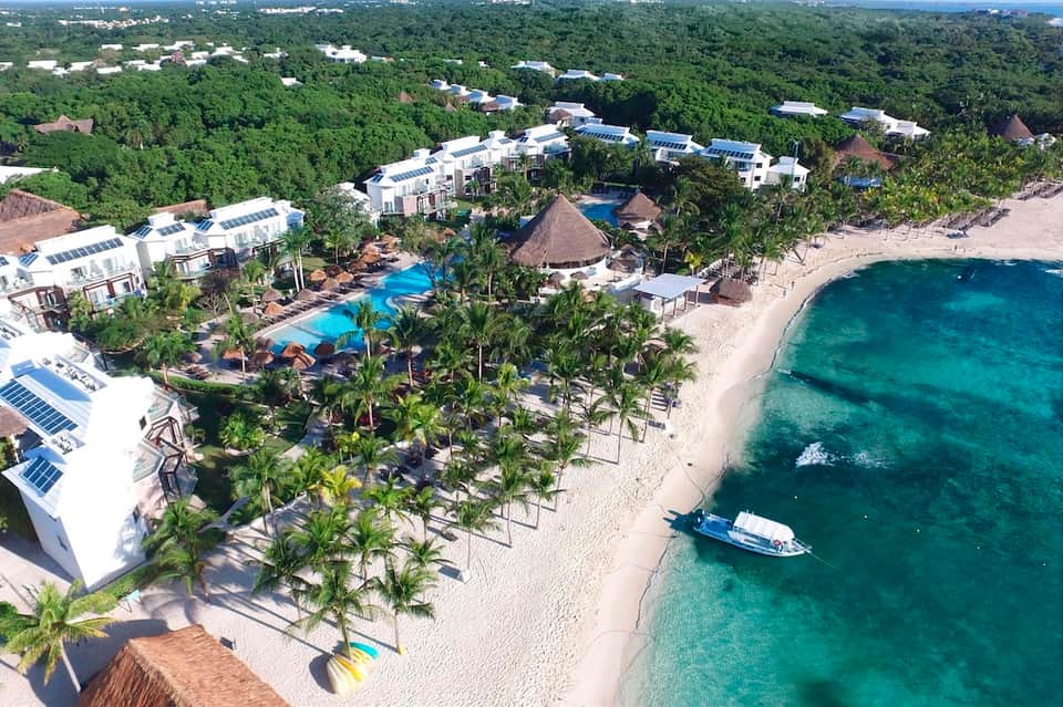 Aerial view of beachfront resort with pool, palm trees, white buildings with solar panels, and turquoise ocean