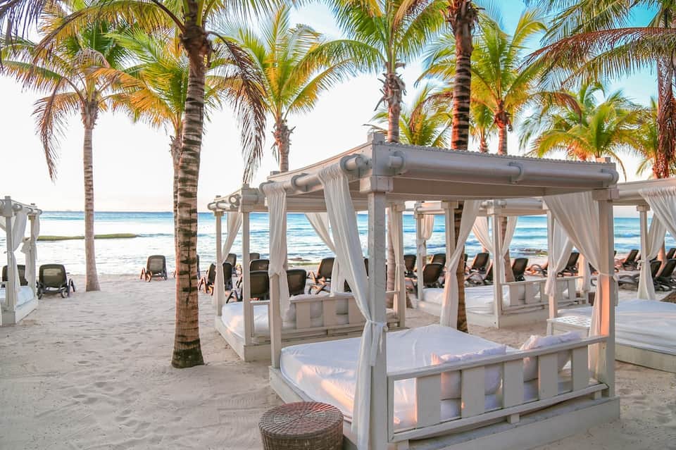 Beach cabanas with white curtains under palm trees overlooking turquoise ocean