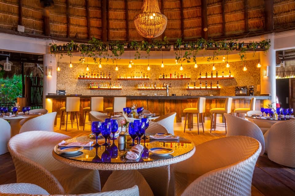 Upscale dining venue with thatched ceiling, wooden bar, hanging greenery, and elegant table settings