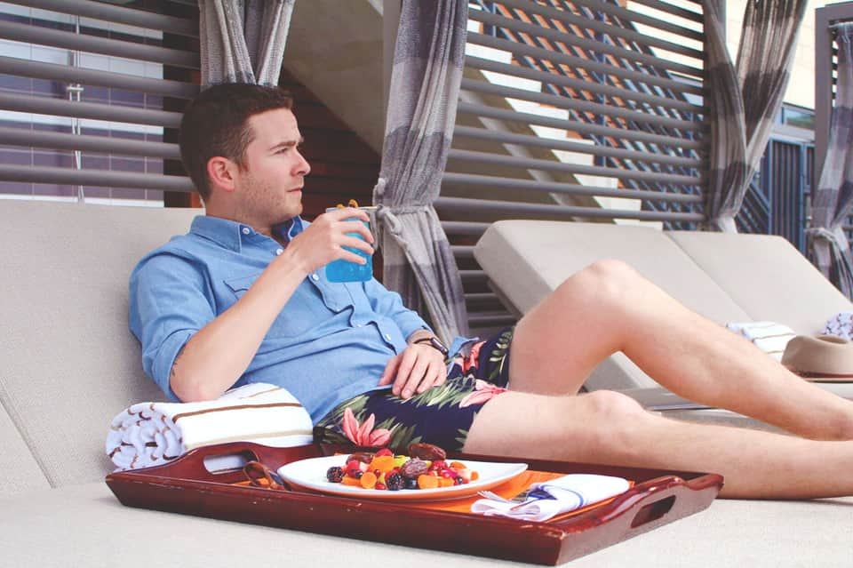 Man in blue shirt relaxing on couch with fruit platter and city view through slatted windows