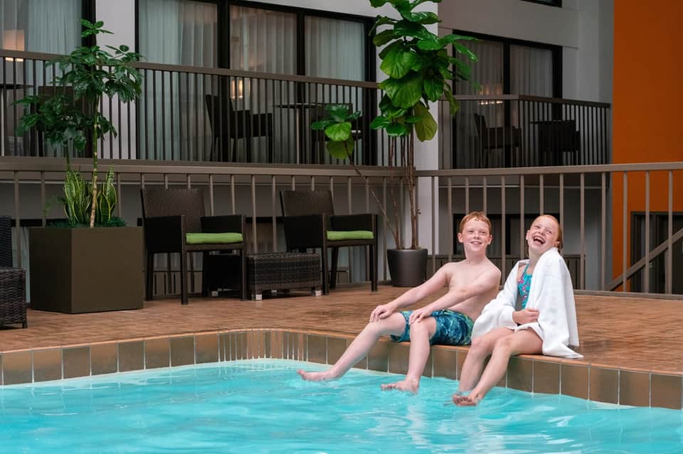 Two children sitting poolside on wooden deck with modern hotel balconies and green accent walls visible