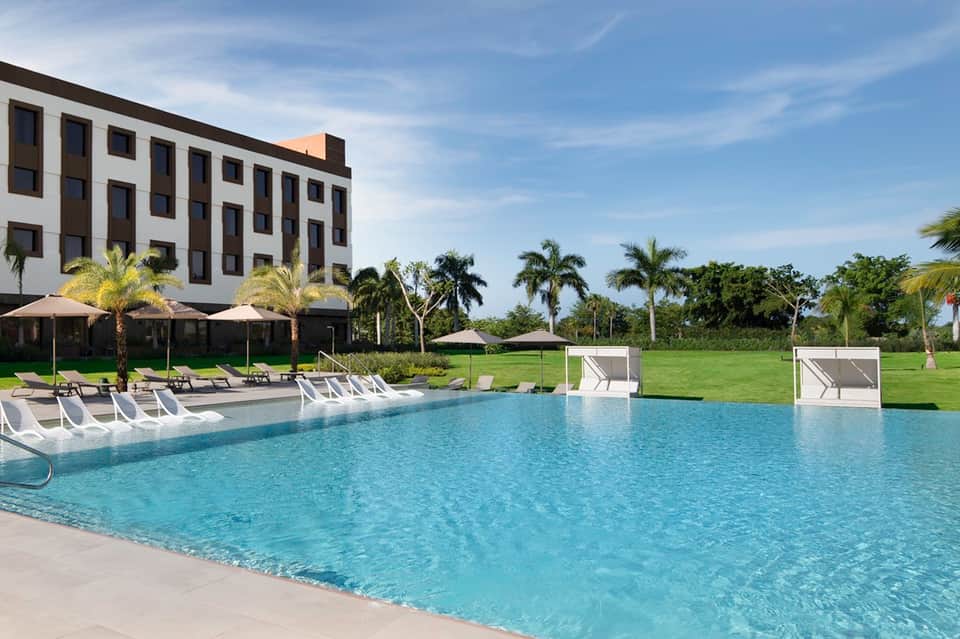 Resort swimming pool with white lounge chairs, umbrellas, palm landscaping, and modern building in background
