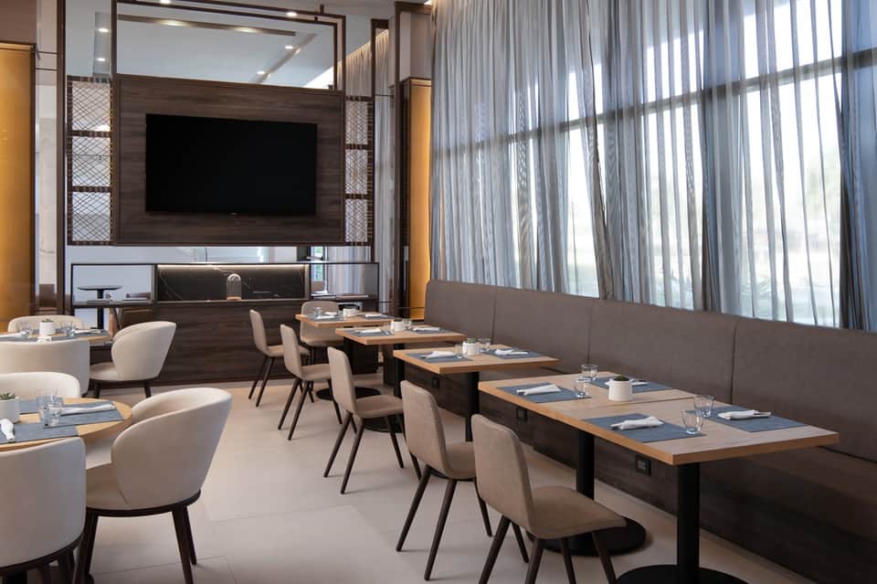Contemporary restaurant interior with wood wall panel, flat-screen TV, white chairs, dining tables, and sheer curtains