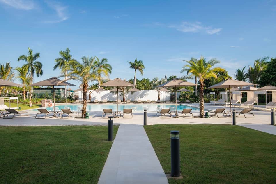 Resort pool area with palm trees, lounge chairs, umbrellas, and manicured lawn