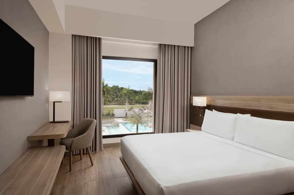 Modern hotel room with king bed, sliding glass doors overlooking pool and palm trees, work desk, and flat-screen TV