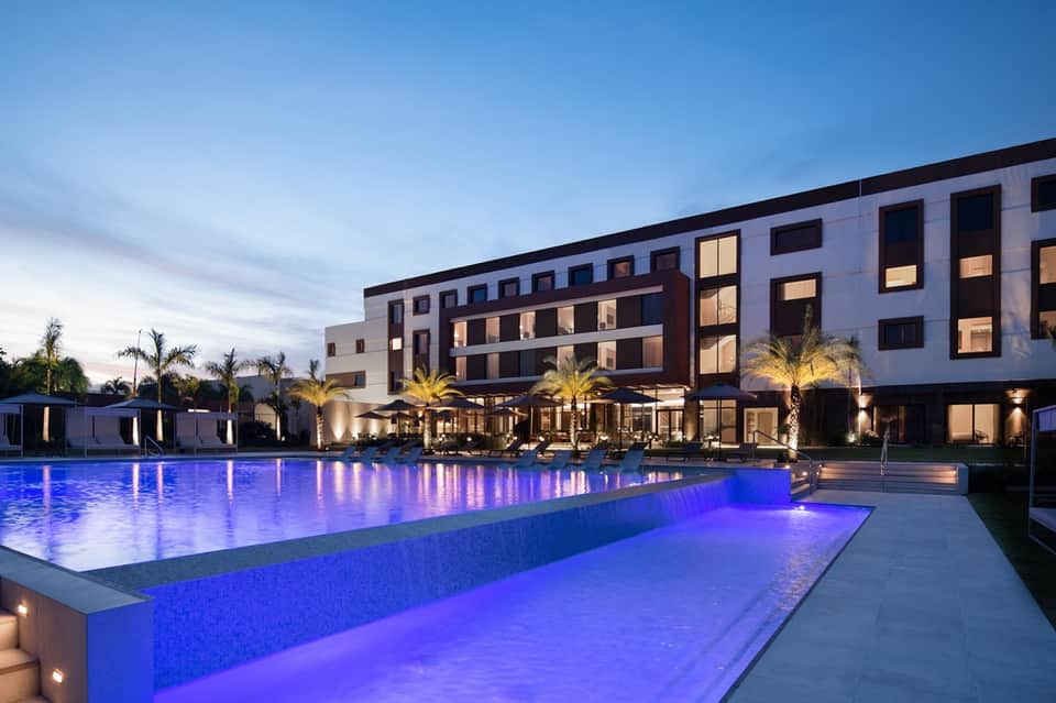 Luxury resort pool illuminated at dusk with modern hotel building, palm trees, and lounge cabanas