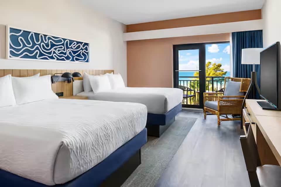 Oceanview hotel room with two beds, coastal artwork, private balcony, and wicker chair