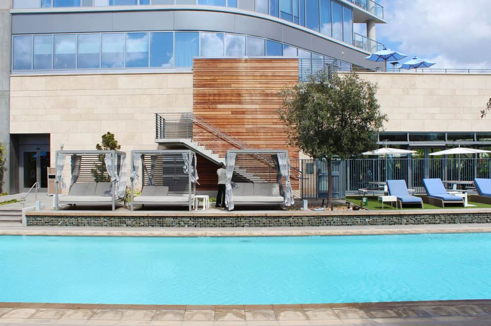 Modern resort pool area with turquoise water, lounge chairs, wooden cabanas, and contemporary building with large windows