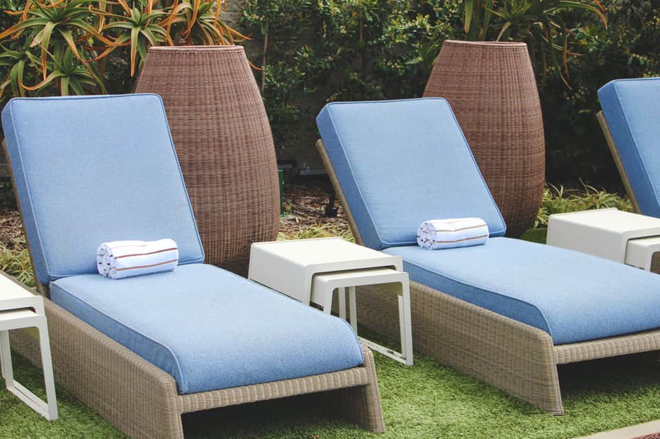 Wicker chaise lounges with blue cushions and rolled towels by landscaped garden