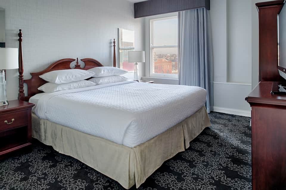 Upscale hotel bedroom with queen bed, burgundy headboard, floor-to-ceiling windows, and city view